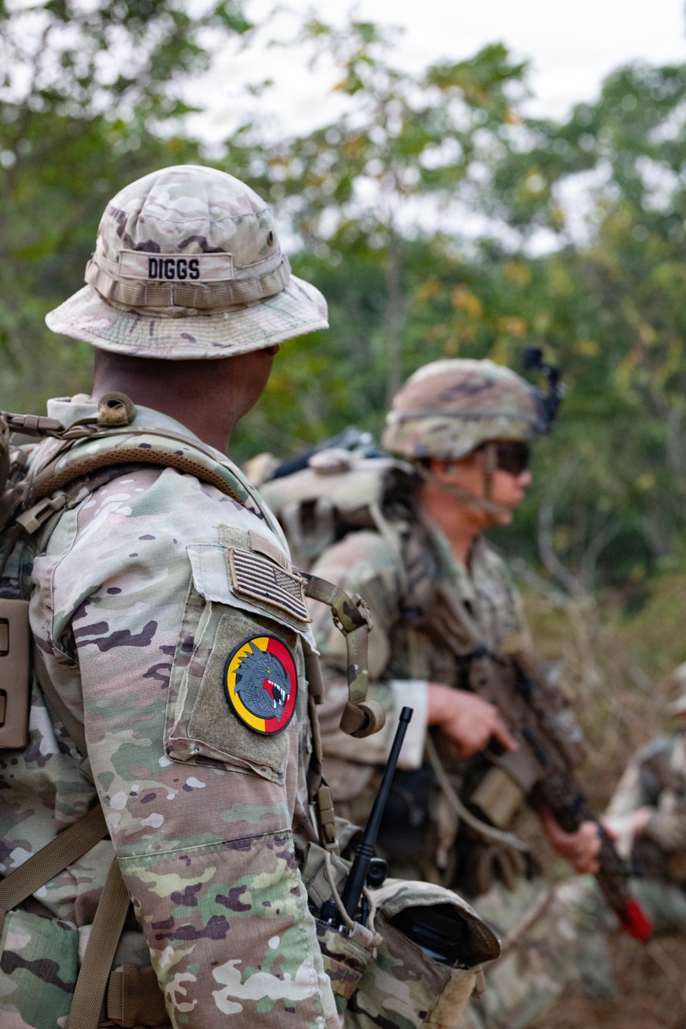 Military personnel in MultiCam/OCP camouflage uniforms are conducting a patrol or field exercise in a wooded outdoor environment. The foreground soldier, identified by name tape as 'DIGGS,' wears a boonie hat and displays a circular unit patch with a wolf/canine head on a red and yellow background on the left sleeve. Multiple soldiers are visible carrying rifles and tactical gear.