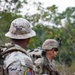 Military personnel in MultiCam/OCP camouflage uniforms are conducting a patrol or field exercise in a wooded outdoor environment. The foreground soldier, identified by name tape as 'DIGGS,' wears a boonie hat and displays a circular unit patch with a wolf/canine head on a red and yellow background on the left sleeve. Multiple soldiers are visible carrying rifles and tactical gear.