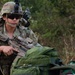 A U.S. Army soldier in full combat gear, including MultiCam (OCP) uniform, tactical helmet with night vision mount, and ballistic sunglasses, holds a rifle with optic attachment. A second individual in a different camouflage pattern (green/black) is partially visible in the foreground right. The setting appears to be an outdoor field training or joint exercise environment.