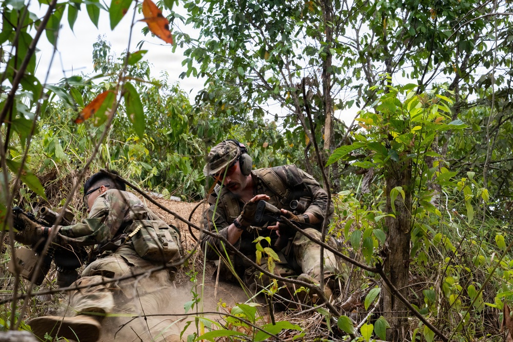 Two soldiers in camouflage uniforms are positioned low in dense jungle or tropical vegetation, appearing to be in a tactical prone/crouching position. One soldier is lying prone while the other is crouching beside them, both holding rifles. The setting suggests a military training exercise or field operation in a tropical environment.