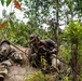 Two soldiers in camouflage uniforms are positioned low in dense jungle or tropical vegetation, appearing to be in a tactical prone/crouching position. One soldier is lying prone while the other is crouching beside them, both holding rifles. The setting suggests a military training exercise or field operation in a tropical environment.