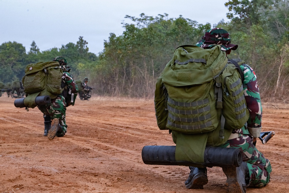The image shows military personnel in camouflage uniforms moving across a red dirt terrain during what appears to be a field exercise or training operation. Soldiers are carrying large tactical backpacks and sleeping mats, advancing along an open dirt track flanked by dry grass and trees. The camouflage pattern and equipment suggest these are likely Indonesian Army (TNI-AD) soldiers based on the distinctive green-brown-black camouflage pattern.