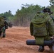 The image shows military personnel in camouflage uniforms moving across a red dirt terrain during what appears to be a field exercise or training operation. Soldiers are carrying large tactical backpacks and sleeping mats, advancing along an open dirt track flanked by dry grass and trees. The camouflage pattern and equipment suggest these are likely Indonesian Army (TNI-AD) soldiers based on the distinctive green-brown-black camouflage pattern.