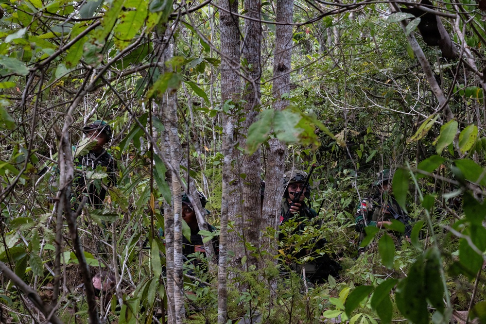 The image shows multiple individuals in military or paramilitary-style camouflage uniforms moving through dense tropical jungle vegetation. At least one person is visibly using a handheld radio or communication device. The scene suggests a patrol, reconnaissance, or field operation in a heavily forested environment.