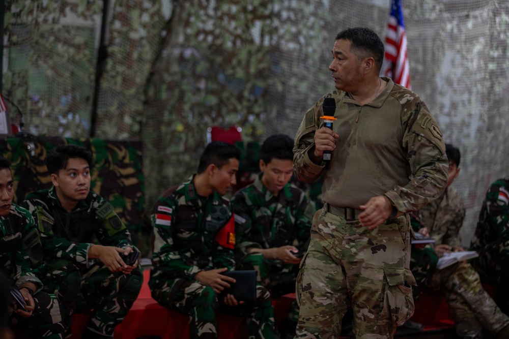 A soldier in US Army MultiCam/OCP camouflage uniform is standing and speaking into a handheld microphone, addressing a group of seated soldiers wearing Indonesian Armed Forces (TNI) woodland camouflage uniforms. An American flag is partially visible in the background, suggesting this is a joint military briefing or training event. The setting appears to be an outdoor or semi-covered military venue with camouflage netting overhead.