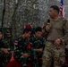 A soldier in US Army MultiCam/OCP camouflage uniform is standing and speaking into a handheld microphone, addressing a group of seated soldiers wearing Indonesian Armed Forces (TNI) woodland camouflage uniforms. An American flag is partially visible in the background, suggesting this is a joint military briefing or training event. The setting appears to be an outdoor or semi-covered military venue with camouflage netting overhead.