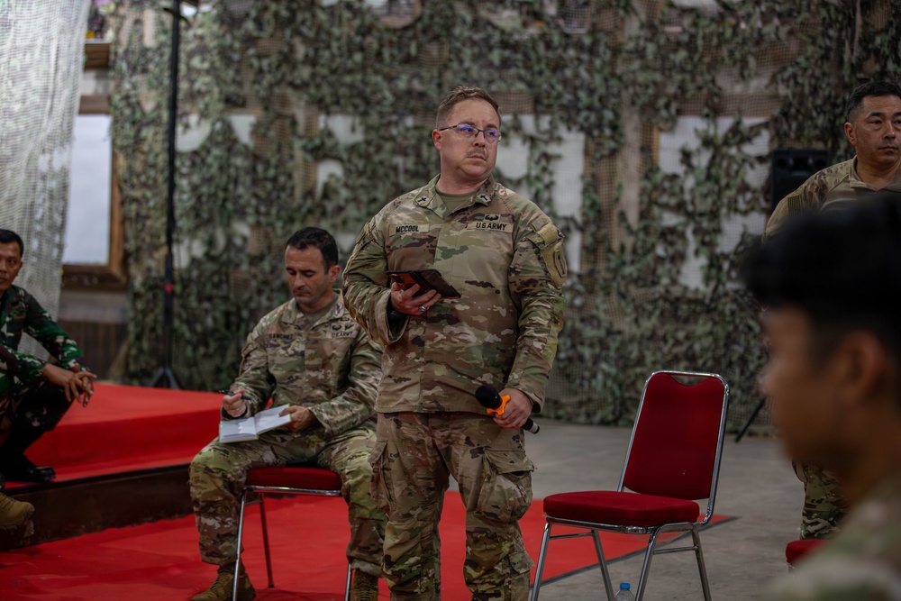 A U.S. Army soldier named McCool stands holding a tablet and microphone in what appears to be a military briefing or ceremony setting. The venue is decorated with camouflage netting as a backdrop, with red carpet and chairs arranged for an audience. Personnel from multiple military forces, including what appears to be Indonesian Army soldiers, are present in the room.