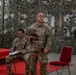 A U.S. Army soldier named McCool stands holding a tablet and microphone in what appears to be a military briefing or ceremony setting. The venue is decorated with camouflage netting as a backdrop, with red carpet and chairs arranged for an audience. Personnel from multiple military forces, including what appears to be Indonesian Army soldiers, are present in the room.