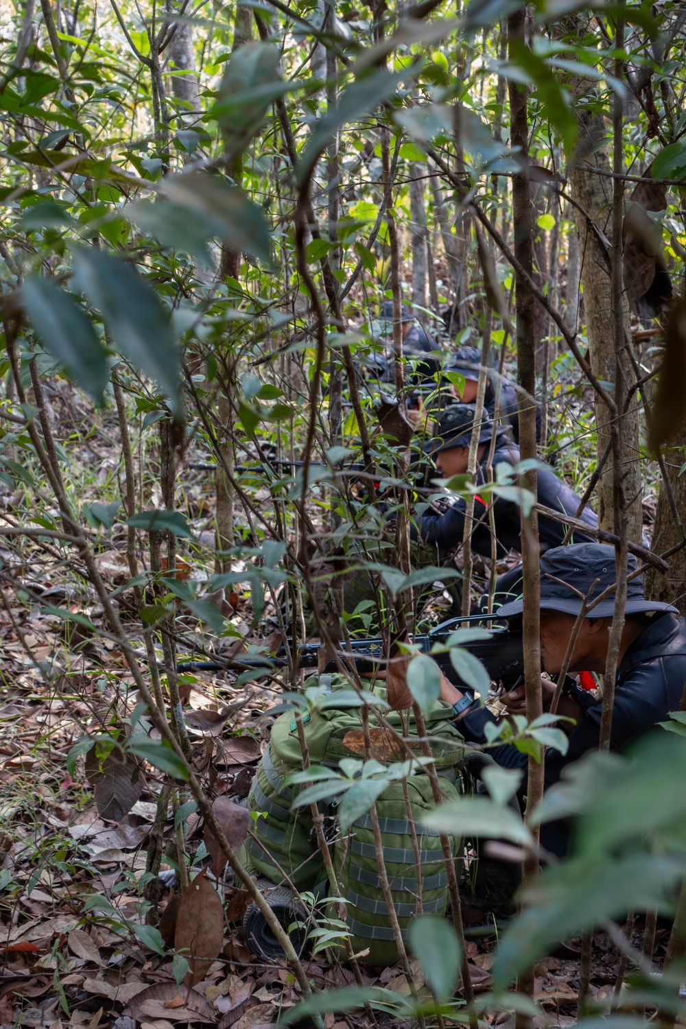 The image shows multiple individuals concealed within dense jungle undergrowth, wearing dark clothing and wide-brimmed hats. They appear to be in a low crouching or prone position among trees and foliage. Firearms and tactical gear, including a green backpack, are visible among the group.