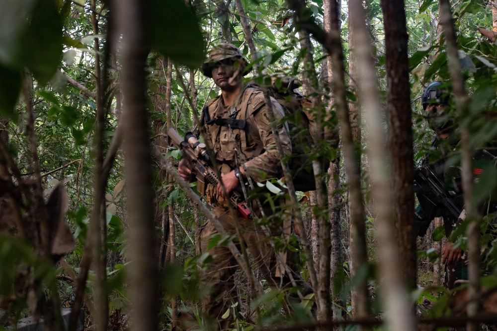 The image shows at least two armed military personnel moving through dense jungle or forest terrain. The primary figure in the foreground wears multicam/OCP camouflage uniform, a boonie hat, and carries a rifle with visible accessories. A second individual is partially visible in the background wearing different camouflage, suggesting a possible joint or multinational military exercise.