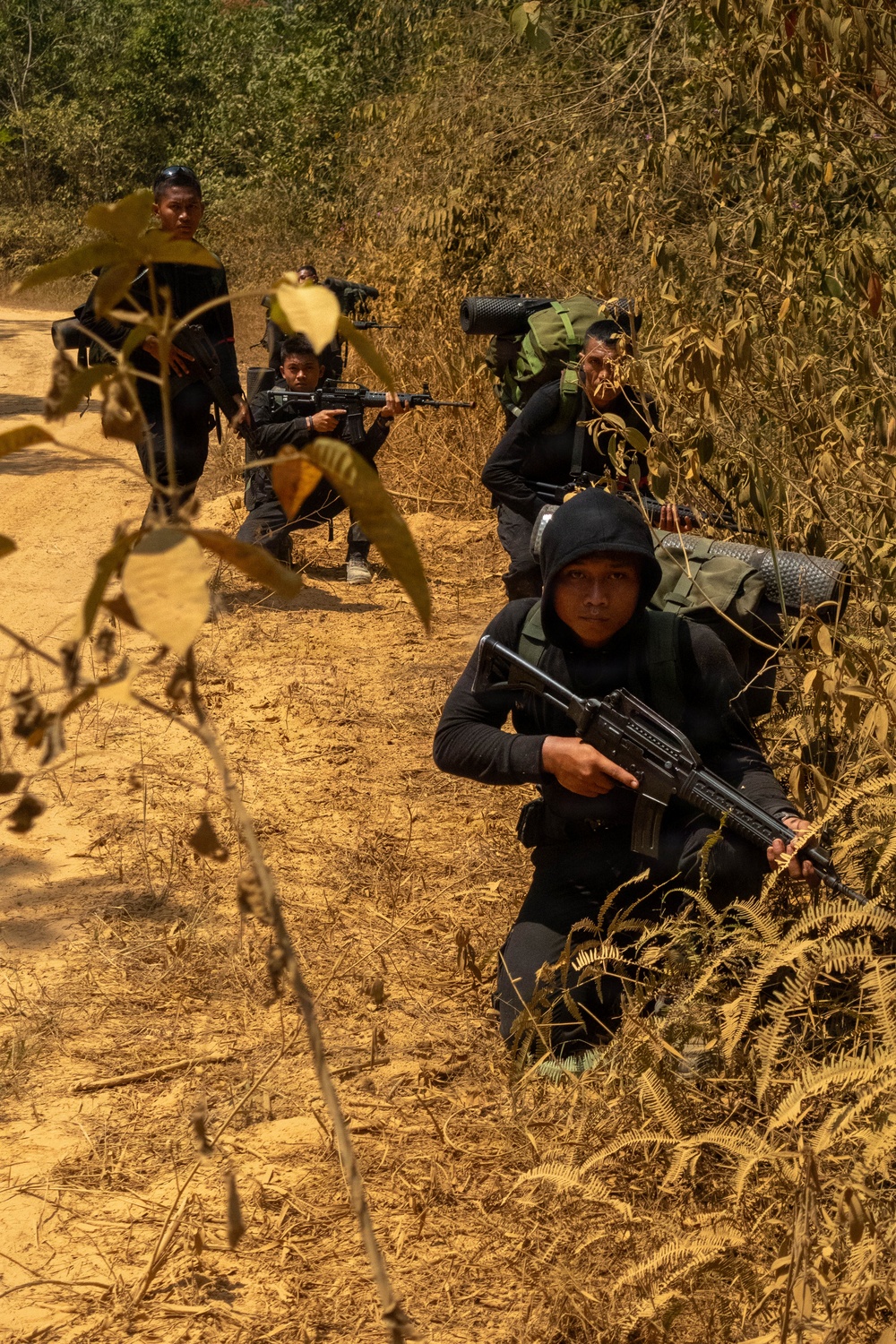 The image shows a group of armed individuals dressed in all-black clothing positioned along the edge of a dirt trail in a dry forest or jungle environment. They appear to be in tactical crouching positions, holding rifles and carrying large backpacks. The scene suggests a patrol, ambush drill, or armed movement through a rural woodland area.