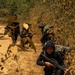 The image shows a group of armed individuals dressed in all-black clothing positioned along the edge of a dirt trail in a dry forest or jungle environment. They appear to be in tactical crouching positions, holding rifles and carrying large backpacks. The scene suggests a patrol, ambush drill, or armed movement through a rural woodland area.