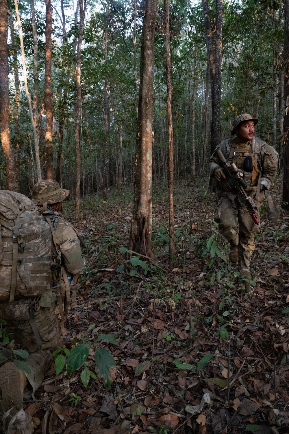 Two soldiers in multicam/camouflage uniforms are conducting a patrol or movement through a tropical or subtropical forest with tall, sparse trees and a leaf-covered ground. One soldier faces away carrying a large rucksack while the other stands alert holding a rifle and scanning the surroundings. The environment suggests a jungle training exercise or operational patrol.