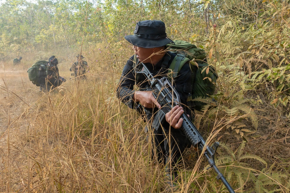 The image shows armed individuals dressed in all-black uniforms moving through dry grassland and sparse woodland terrain. The primary subject in the foreground is crouching in tall dry grass while holding a rifle, with at least two additional similarly equipped individuals visible in the background. The setting appears to be a tropical or subtropical environment during a dry season.