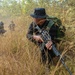 The image shows armed individuals dressed in all-black uniforms moving through dry grassland and sparse woodland terrain. The primary subject in the foreground is crouching in tall dry grass while holding a rifle, with at least two additional similarly equipped individuals visible in the background. The setting appears to be a tropical or subtropical environment during a dry season.