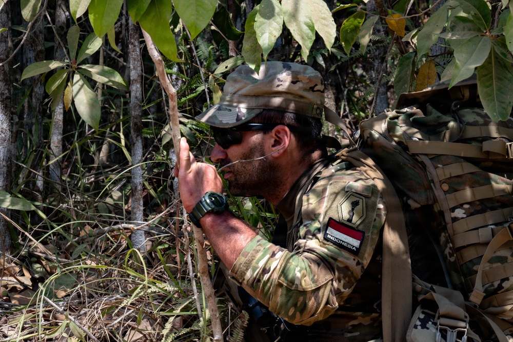 A soldier wearing Indonesian military insignia is crouched in a jungle environment, appearing to be in a tactical observation or patrol position. He is equipped with full combat gear including a multicam camouflage uniform, tactical backpack, and cap. The setting suggests a jungle warfare training exercise or field operation.