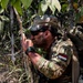A soldier wearing Indonesian military insignia is crouched in a jungle environment, appearing to be in a tactical observation or patrol position. He is equipped with full combat gear including a multicam camouflage uniform, tactical backpack, and cap. The setting suggests a jungle warfare training exercise or field operation.