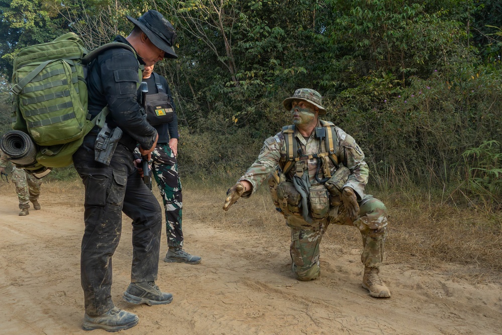 The image shows what appears to be a military training exercise in a forested, sandy terrain environment. A soldier in full camouflage with face paint is kneeling and gesturing/giving instructions to another individual wearing tactical gear and a large rucksack. A third person in mixed military attire is visible in the background.
