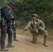 The image shows what appears to be a military training exercise in a forested, sandy terrain environment. A soldier in full camouflage with face paint is kneeling and gesturing/giving instructions to another individual wearing tactical gear and a large rucksack. A third person in mixed military attire is visible in the background.
