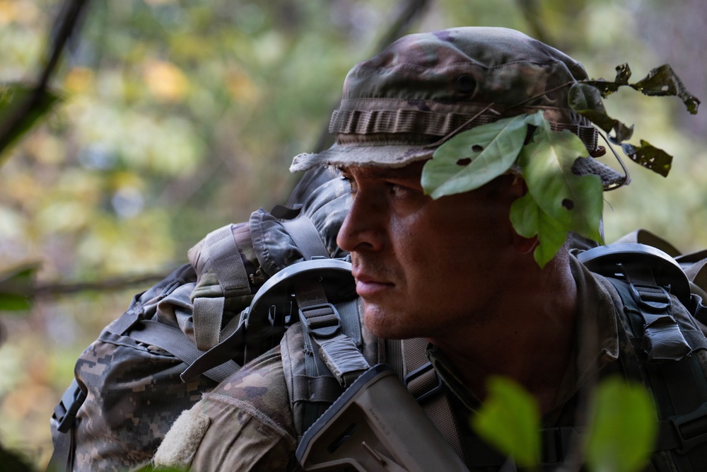 A soldier in camouflage uniform and boonie hat with foliage attached is photographed in a wooded environment. The individual is wearing a large tactical rucksack and appears to be on patrol or conducting a field training exercise. The close-up portrait captures a focused, alert expression against a blurred forest background.