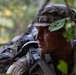 A soldier in camouflage uniform and boonie hat with foliage attached is photographed in a wooded environment. The individual is wearing a large tactical rucksack and appears to be on patrol or conducting a field training exercise. The close-up portrait captures a focused, alert expression against a blurred forest background.