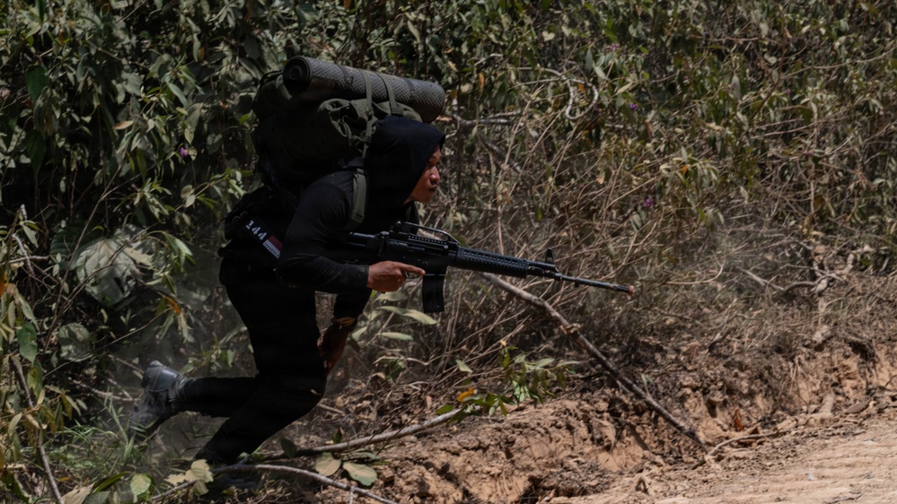 A person dressed in all-black tactical clothing and carrying a large military-style backpack is moving through dense vegetation along a dirt path or embankment. They are holding what appears to be a semi-automatic rifle in a forward-facing position. The setting appears to be a jungle or forested environment under bright daylight conditions.