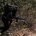 A person dressed in all-black tactical clothing and carrying a large military-style backpack is moving through dense vegetation along a dirt path or embankment. They are holding what appears to be a semi-automatic rifle in a forward-facing position. The setting appears to be a jungle or forested environment under bright daylight conditions.