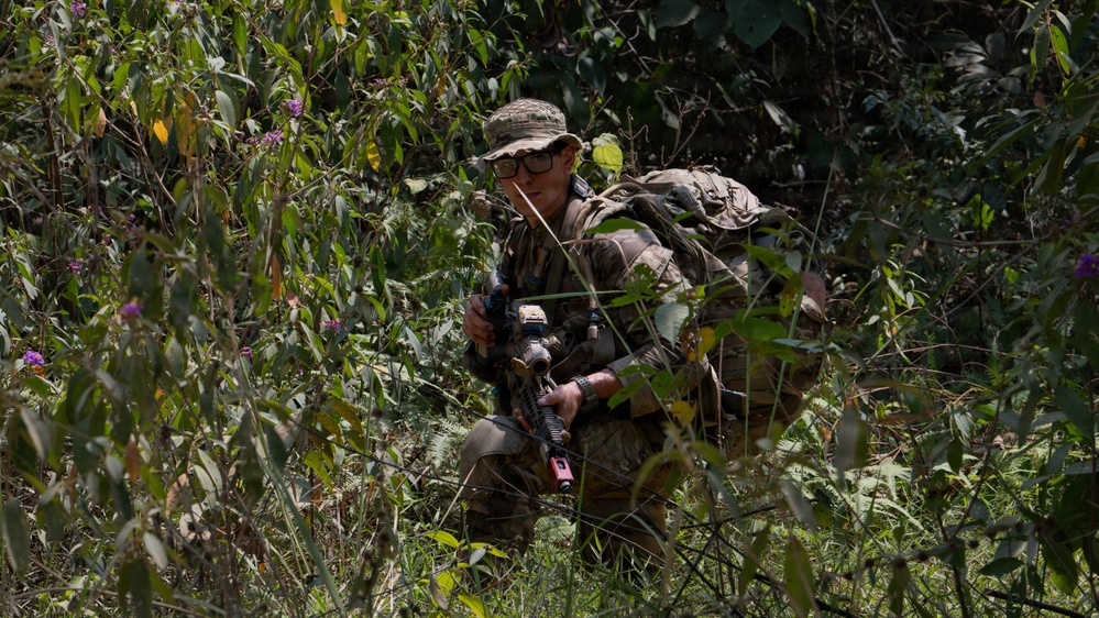 A soldier in full camouflage gear is crouching in dense vegetation, appearing to move stealthily through a jungle or forested environment. The individual is equipped with a rifle featuring tactical accessories and is wearing a boonie hat and protective eyewear. The scene suggests a military field exercise or patrol operation.