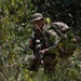 A soldier in full camouflage gear is crouching in dense vegetation, appearing to move stealthily through a jungle or forested environment. The individual is equipped with a rifle featuring tactical accessories and is wearing a boonie hat and protective eyewear. The scene suggests a military field exercise or patrol operation.