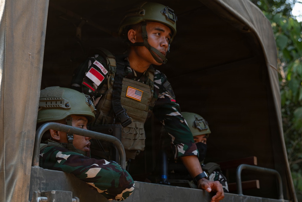 Three military personnel are positioned in the back of a covered military transport vehicle. The standing soldier prominently displays both an Indonesian flag patch and a US flag patch on his tactical vest, suggesting a joint military exercise or operation. All personnel are wearing camouflage uniforms and helmets consistent with Indonesian Armed Forces (TNI) pattern.
