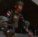 Three military personnel are positioned in the back of a covered military transport vehicle. The standing soldier prominently displays both an Indonesian flag patch and a US flag patch on his tactical vest, suggesting a joint military exercise or operation. All personnel are wearing camouflage uniforms and helmets consistent with Indonesian Armed Forces (TNI) pattern.