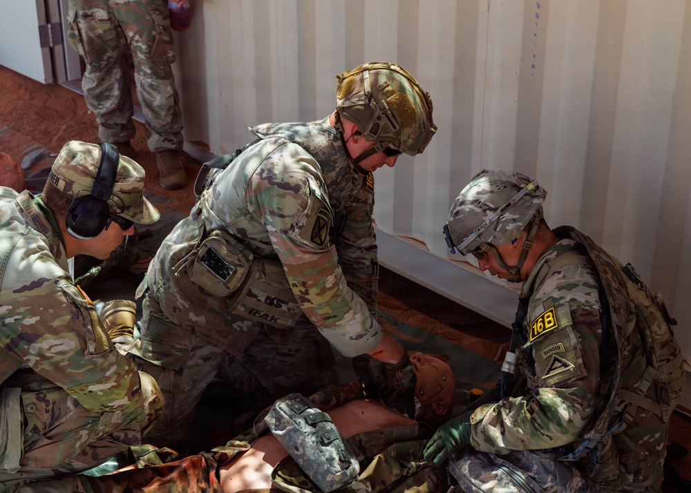 Several U.S. Army soldiers in OCP (Operational Camouflage Pattern) uniforms are conducting a tactical combat casualty care (TCCC) training exercise. They are attending to what appears to be a training mannequin or simulated casualty on the ground near a metal shipping container. At least one soldier wears a tactical helmet with NVG mount and another displays a '16B' unit patch with a Ranger tab.