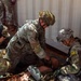 Several U.S. Army soldiers in OCP (Operational Camouflage Pattern) uniforms are conducting a tactical combat casualty care (TCCC) training exercise. They are attending to what appears to be a training mannequin or simulated casualty on the ground near a metal shipping container. At least one soldier wears a tactical helmet with NVG mount and another displays a '16B' unit patch with a Ranger tab.