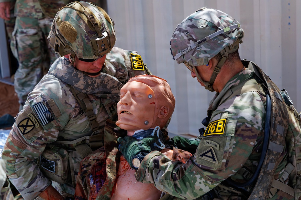Two U.S. Army soldiers in full combat gear are conducting a tactical combat casualty care (TCCC) training exercise using a medical simulation mannequin. The soldiers wear multicam uniforms with unit patches including 10th Mountain Division and Ranger tab, and are labeled with yellow competition or evaluation numbers (16A and 16B). The mannequin simulates a wounded casualty with visible simulated blood and a cervical collar applied.