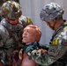 Two U.S. Army soldiers in full combat gear are conducting a tactical combat casualty care (TCCC) training exercise using a medical simulation mannequin. The soldiers wear multicam uniforms with unit patches including 10th Mountain Division and Ranger tab, and are labeled with yellow competition or evaluation numbers (16A and 16B). The mannequin simulates a wounded casualty with visible simulated blood and a cervical collar applied.