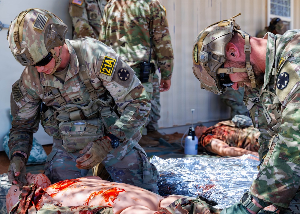 Two U.S. Army soldiers in OCP (Operational Camouflage Pattern) uniforms are performing tactical combat casualty care (TCCC) on a simulated casualty during a medical training exercise. The scenario involves moulage (simulated wounds with artificial blood) on a mannequin or training patient placed on a mylar emergency blanket. Additional personnel in uniform are visible in the background observing or participating in the exercise.