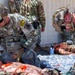 Two U.S. Army soldiers in OCP (Operational Camouflage Pattern) uniforms are performing tactical combat casualty care (TCCC) on a simulated casualty during a medical training exercise. The scenario involves moulage (simulated wounds with artificial blood) on a mannequin or training patient placed on a mylar emergency blanket. Additional personnel in uniform are visible in the background observing or participating in the exercise.