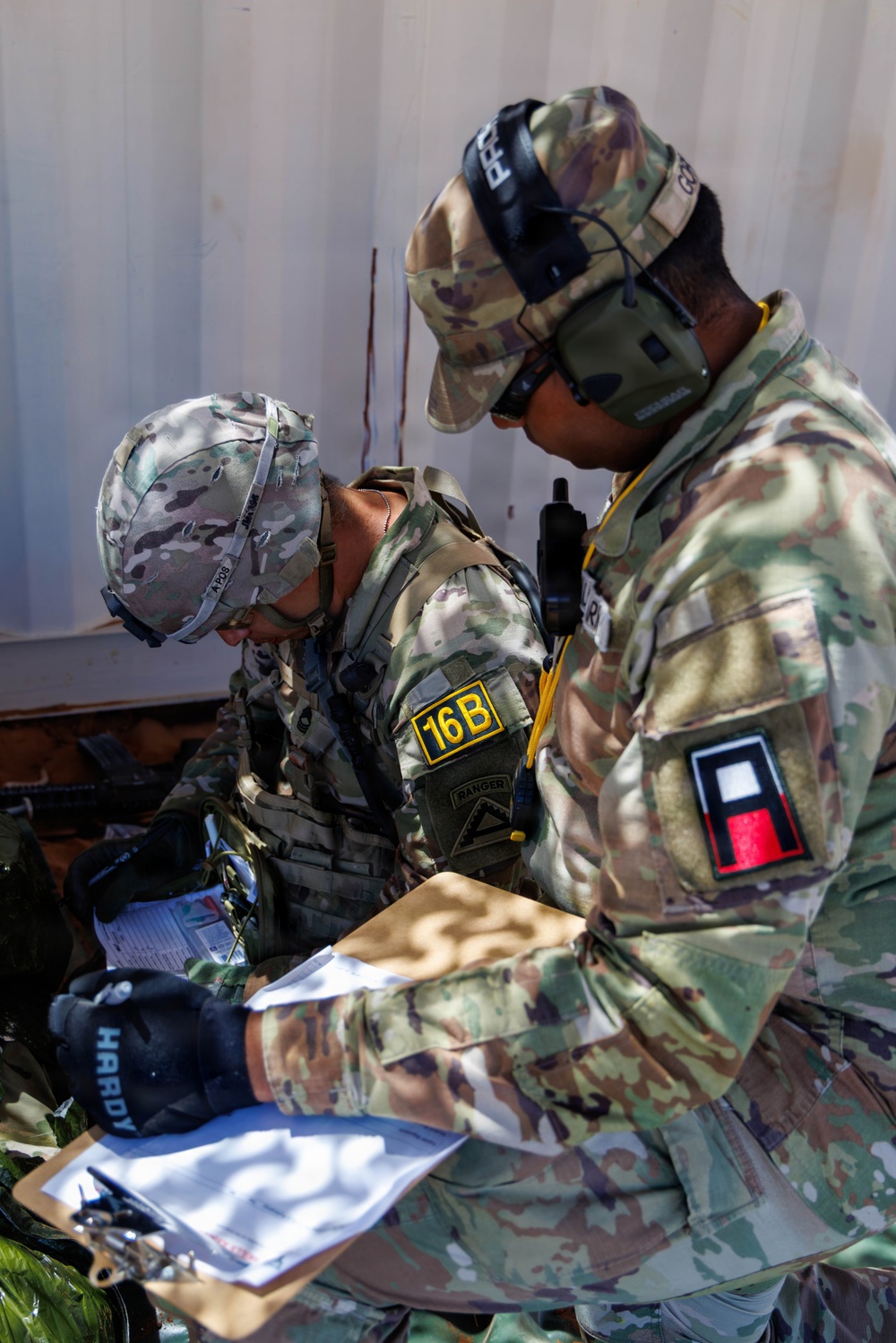 Two U.S. Army soldiers in OCP (Operational Camouflage Pattern) uniforms are reviewing documents on a clipboard, likely during a training exercise or operational briefing. The left soldier wears a '16B' MOS patch (Air Defense Early Warning Systems Operator) and a Ranger tab, while the right soldier wears tactical hearing protection and carries a radio. Both are focused on paperwork near what appears to be a shipping container.