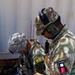 Two U.S. Army soldiers in OCP (Operational Camouflage Pattern) uniforms are reviewing documents on a clipboard, likely during a training exercise or operational briefing. The left soldier wears a '16B' MOS patch (Air Defense Early Warning Systems Operator) and a Ranger tab, while the right soldier wears tactical hearing protection and carries a radio. Both are focused on paperwork near what appears to be a shipping container.