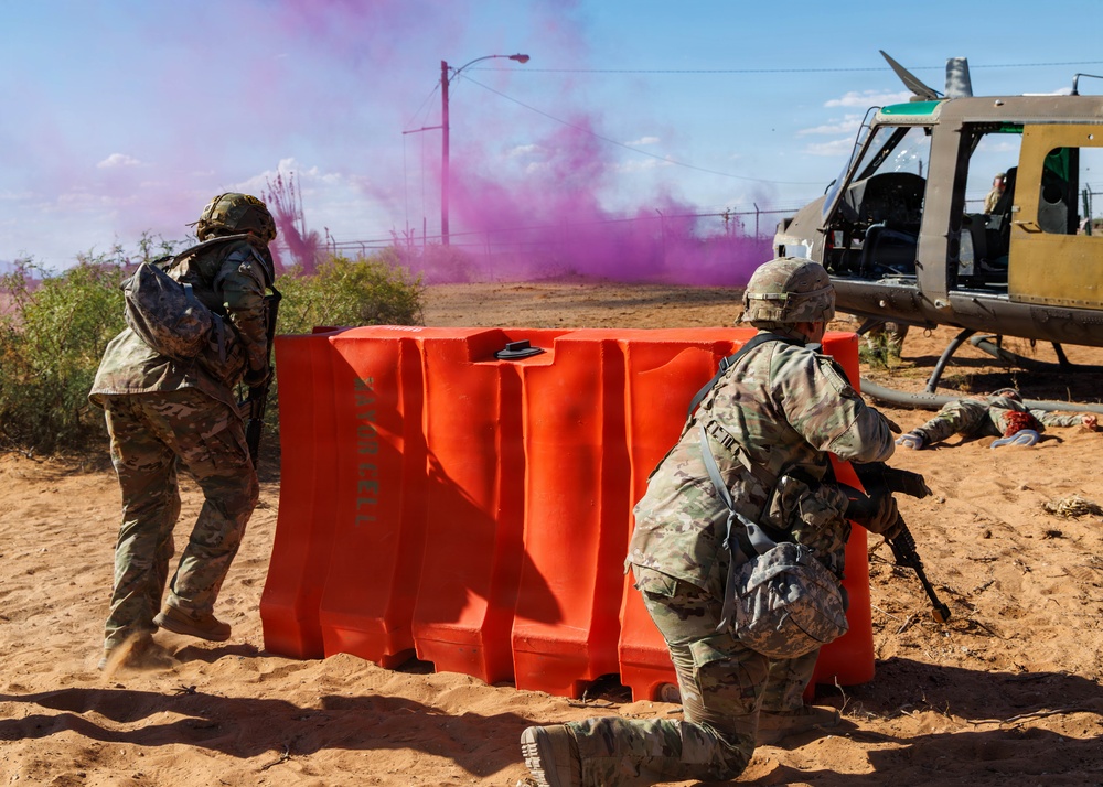 A military training exercise is depicted with two soldiers in camouflage uniforms taking cover behind a large orange barrier. Purple smoke grenades are deployed in the background, and a military helicopter (appears to be a UH-1 Huey variant) is visible on the right side. A simulated casualty figure is visible on the ground near the helicopter, indicating a combat scenario drill.