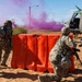 A military training exercise is depicted with two soldiers in camouflage uniforms taking cover behind a large orange barrier. Purple smoke grenades are deployed in the background, and a military helicopter (appears to be a UH-1 Huey variant) is visible on the right side. A simulated casualty figure is visible on the ground near the helicopter, indicating a combat scenario drill.