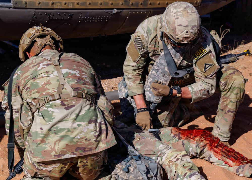 Two soldiers in MultiCam/OCP camouflage uniforms are performing tactical combat casualty care (TCCC) on a downed soldier lying on red desert terrain. The soldier identified by name tape 'VOZA' is actively treating what appears to be a simulated leg wound with visible red (simulated blood) on the casualty's lower extremity. This appears to be a training exercise or operational medical response scenario near a military vehicle.