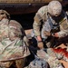 Two soldiers in MultiCam/OCP camouflage uniforms are performing tactical combat casualty care (TCCC) on a downed soldier lying on red desert terrain. The soldier identified by name tape 'VOZA' is actively treating what appears to be a simulated leg wound with visible red (simulated blood) on the casualty's lower extremity. This appears to be a training exercise or operational medical response scenario near a military vehicle.