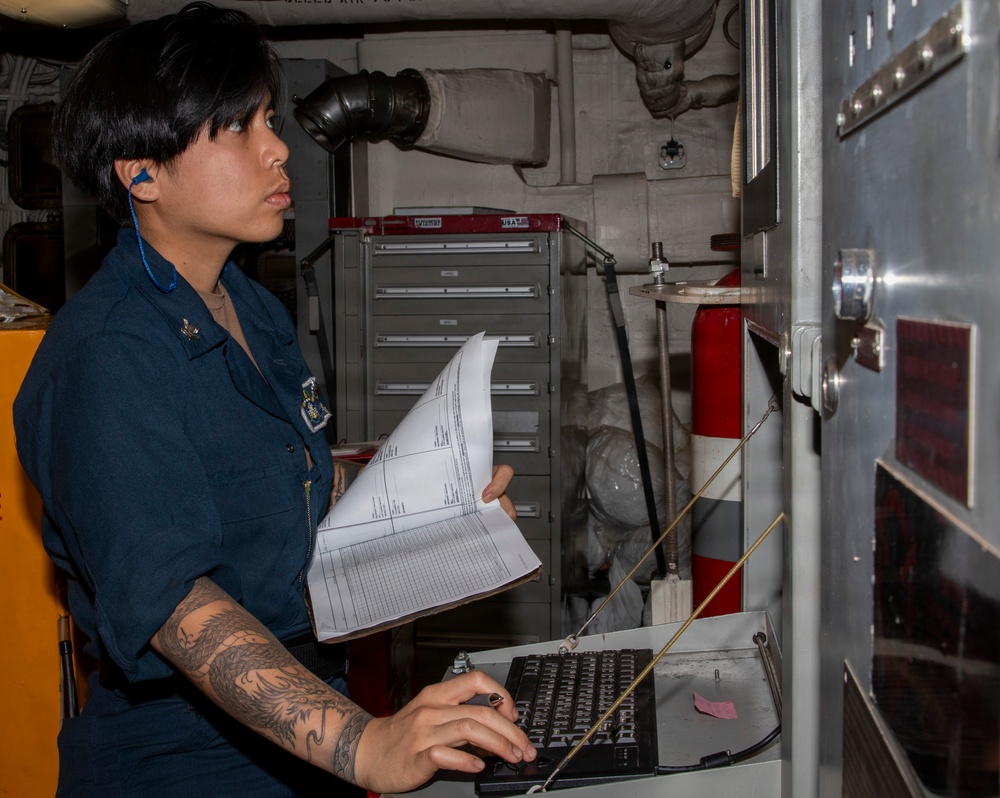 A U.S. Navy sailor in dark blue coveralls is working at a workstation aboard a naval vessel, referencing printed documents while operating a keyboard. The environment appears to be an interior compartment or workspace on a ship, identifiable by the industrial piping, tool chest, and fire extinguisher visible in the background. The sailor is wearing hearing protection (blue earplugs) and has visible tattoos on their right forearm.
