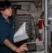 A U.S. Navy sailor in dark blue coveralls is working at a workstation aboard a naval vessel, referencing printed documents while operating a keyboard. The environment appears to be an interior compartment or workspace on a ship, identifiable by the industrial piping, tool chest, and fire extinguisher visible in the background. The sailor is wearing hearing protection (blue earplugs) and has visible tattoos on their right forearm.