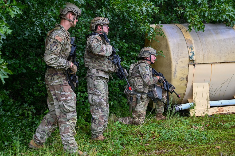 Three military personnel in MultiCam/OCP camouflage uniforms are conducting what appears to be a tactical training exercise in a vegetated area. They are positioned near a large cylindrical metal tank or barrel, with at least one soldier in a crouched position appearing to call out or communicate. All three are armed with rifles and wearing tactical helmets and body armor.