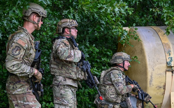 Three military personnel in MultiCam/OCP camouflage uniforms are conducting what appears to be a tactical training exercise in a vegetated area. They are positioned near a large cylindrical metal tank or barrel, with at least one soldier in a crouched position appearing to call out or communicate. All three are armed with rifles and wearing tactical helmets and body armor.