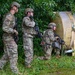 Three military personnel in MultiCam/OCP camouflage uniforms are conducting what appears to be a tactical training exercise in a vegetated area. They are positioned near a large cylindrical metal tank or barrel, with at least one soldier in a crouched position appearing to call out or communicate. All three are armed with rifles and wearing tactical helmets and body armor.