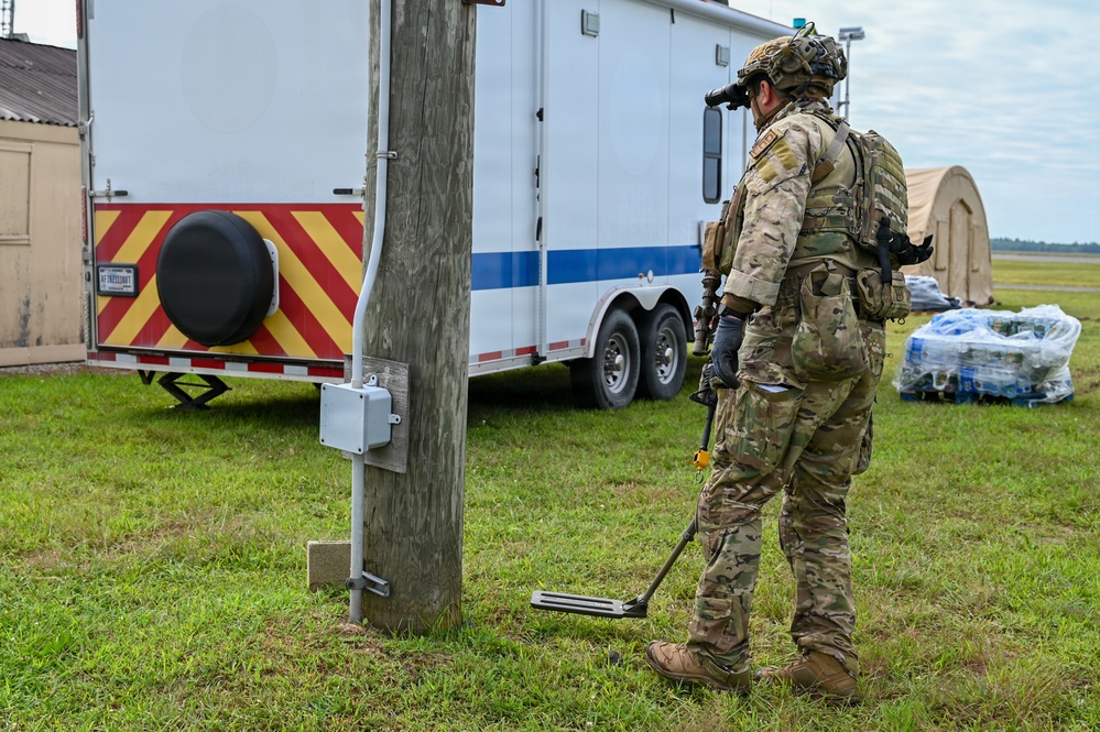 A fully equipped military or law enforcement operator in multicam camouflage is using a metal detector or ground-scanning device near a utility pole on a grassy area. The scene appears to be a forward operating base or staging area, with a white and red/yellow chevron-marked trailer, a tan shelter structure, and palletized water supplies visible in the background. The activity suggests an explosive ordnance disposal (EOD), route clearance, or security sweep operation.