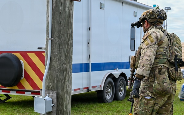 A fully equipped military or law enforcement operator in multicam camouflage is using a metal detector or ground-scanning device near a utility pole on a grassy area. The scene appears to be a forward operating base or staging area, with a white and red/yellow chevron-marked trailer, a tan shelter structure, and palletized water supplies visible in the background. The activity suggests an explosive ordnance disposal (EOD), route clearance, or security sweep operation.